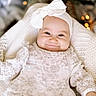 baby, infant, child, smiling, portrait, indoor, christmas, holiday, bow, headband, white_clothing, floral_clothing, blanket, cushion, sitting, lying_down, bokeh, warm_lighting, festive, cozy