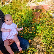 Meredith is registered to the contest to win money with this photo: baby, child, infant, parent, adult, garden, flowers, orange_flowers, outdoor, nature, sitting, holding, portrait, smiling, pink_dress, clothing, sunlight, plants, rocks, family