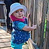 toddler, child, hat, blue_clothing, vest, wooden_deck, wooden_railing, outdoor, daylight, person, smiling, standing, curious, casual_wear, nature, portrait, footwear, sunlight, hand, background_person