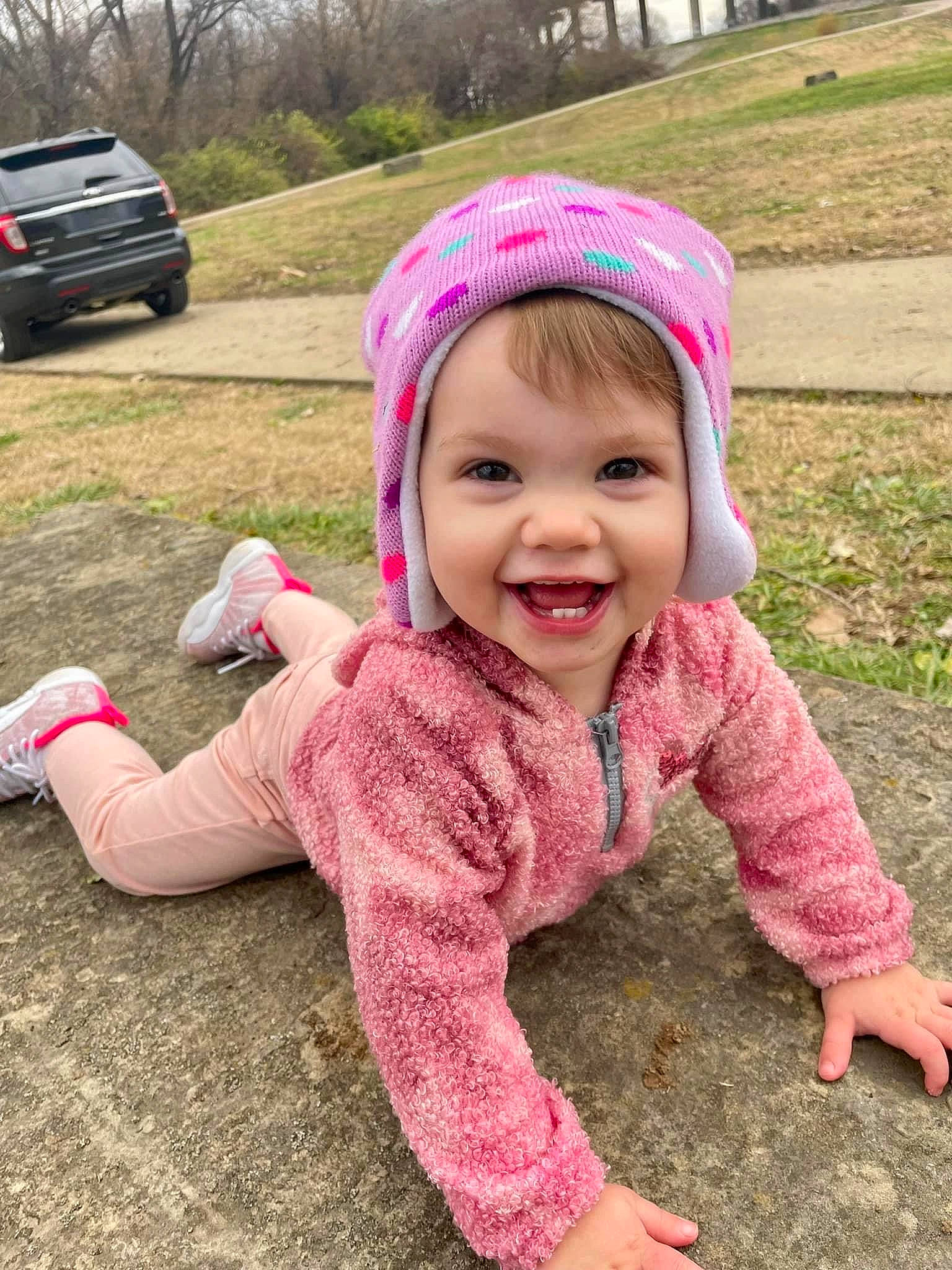 Amaryllis is registered to the contest to win money with this photo: baby_toddler_clothing, cap, car, face, facial_expression, grass, happy, headwear, joy, mammal, outerwear, person, pink, plant, public_space, skin, sleeve, smile, tire, toddler
