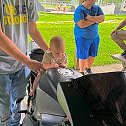 Kaisen joined the competition — help win amazing prizes! baby, motorcycle, adult, child, person, blue_shorts, blue_shirt, gray_shirt, grass, outdoor, fence, wooden_pole, hand, legs, concrete, park, people, casual_clothing, summer, daylight