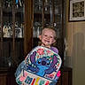 child, girl, smiling, portrait, backpack, stitch, cartoon, school_bag, purple_clothing, indoor, home, display_cabinet, glassware, furniture, wood, framed_picture, standing, happy, colorful, photograph