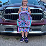 Athena is registered to the contest to win money with this photo: child, girl, smiling, outdoor, pickup_truck, vehicle, backpack, cartoon, stitch, colorful, dress, shoes, standing, portrait, driveway, pavement, garage, sky, daylight, school
