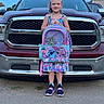 child, girl, smiling, outdoor, pickup_truck, vehicle, backpack, cartoon, stitch, colorful, dress, shoes, standing, portrait, driveway, pavement, garage, sky, daylight, school
