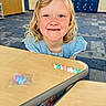 child, smiling, freckles, blonde_hair, blue_dress, portrait, classroom, school, desk, table, toy, miniature_ducks, colorful_toys, carpet, indoor, happy, face, sitting, playful, photography