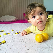 Adam a rejoint le concours — aidez-le/la à gagner de superbes lots ! toddler, child, bed, yellow_clothing, candy, fruit, face, eyes, hair, curious, indoor, pillow, blanket, tablecloth, yellow, lying_down, expression, soft_lighting, room, young_child