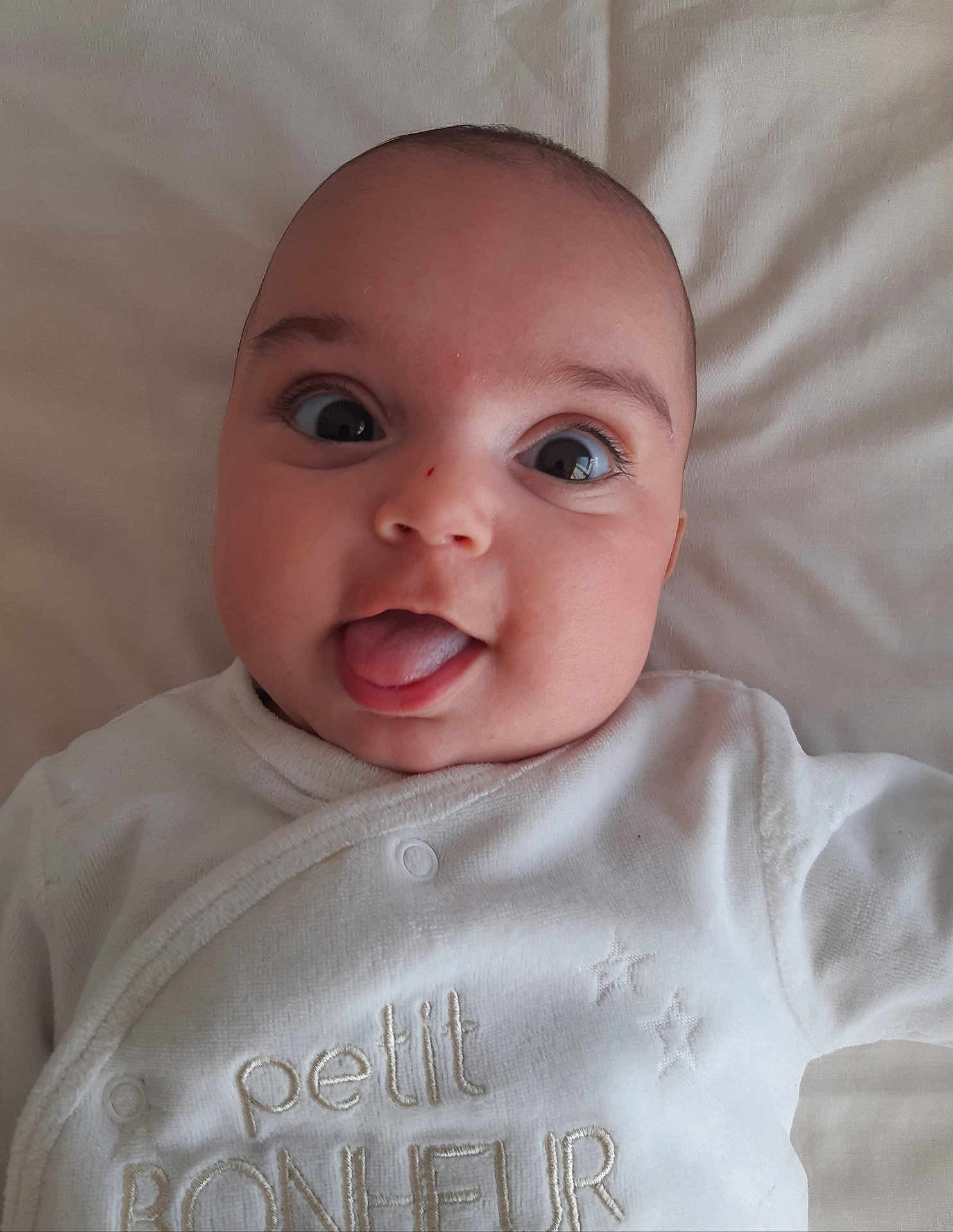 Eliza participe au concours pour gagner de l'argent avec cette photo : baby, infant, face, tongue_out, white_clothing, closeup, cute, smiling, lying_down, soft_texture, cheeks, eyes, head, expression, newborn, child, adorable, portrait, sleepwear, fabric