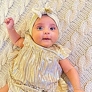 Alayna is registered to the contest to win money with this photo: baby, infant, child, portrait, lying_down, knitted_blanket, golden_dress, headband, bow, wide_eyes, smiling, raised_arm, hands, arms, indoor, cozy, textile, fashion, cute, closeup
