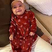 Alayna is registered to the contest to win money with this photo: adult_arm, baby, bow, child, couch, cushions, fabric, face, floral_dress, happy, headband, home, indoor, infant, photography, portrait, red_dress, seated, smiling, white_pants