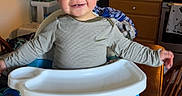 Wyatt is registered to the contest to win money with this photo: booster_seat, casual, child, clothing, face, furniture, happy, high_chair, home, indoor, kitchen, long_sleeve_shirt, oven, pants, person, smiling, sneakers, table, toddler, wooden_cabinet