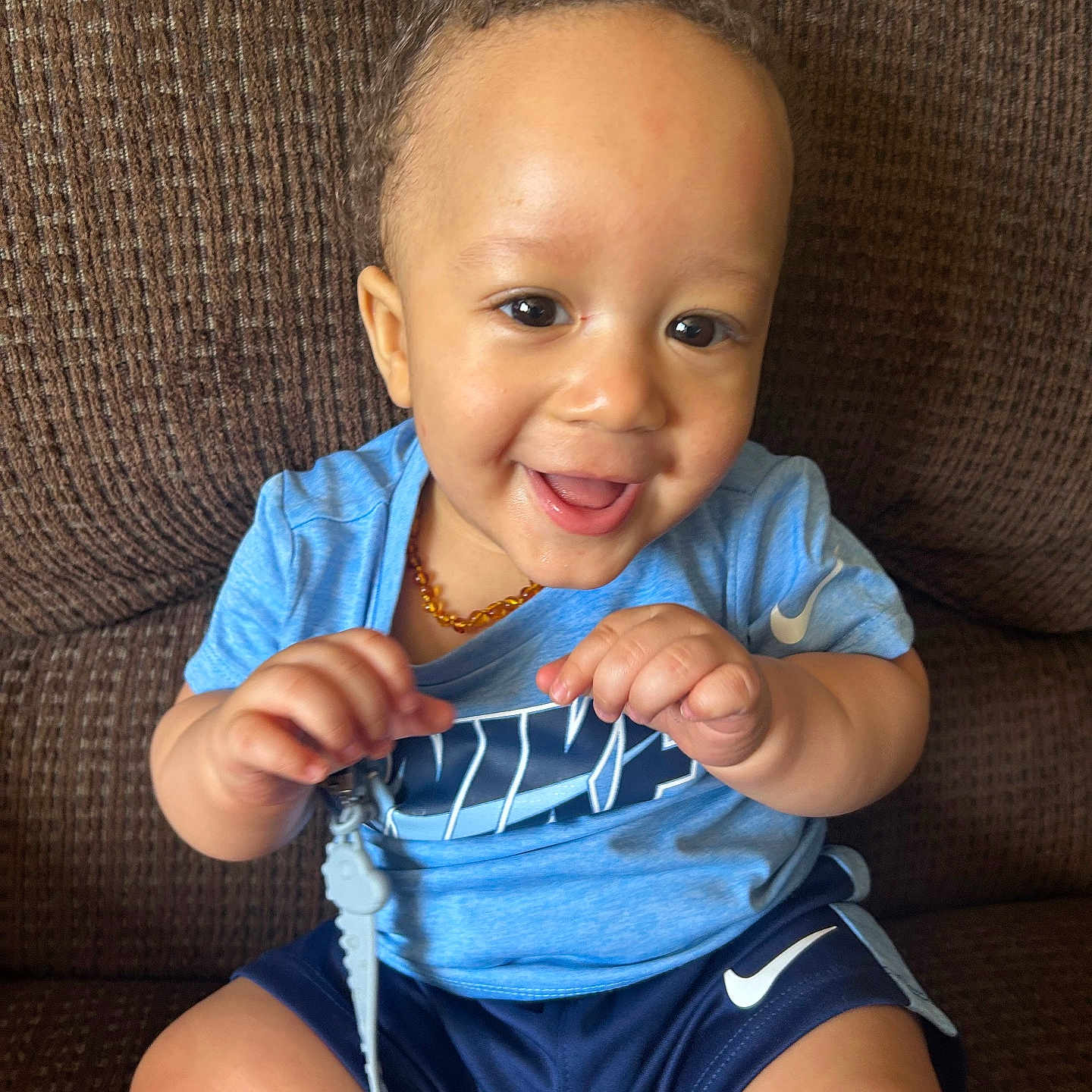 Ajion is registered to the contest to win money with this photo: baby, blue_shirt, child, clip_on, couch, curly_hair, cute, face, hand, happy, indoor, nike, person, playful, portrait, shorts, sitting, smiling, toddler, toy