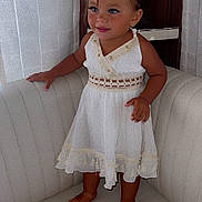 Djaya participe au concours pour gagner de l'argent avec cette photo : toddler, child, barefoot, white_dress, lace, couch, smiling, curly_hair, indoors, furniture, curtains, wooden_background, cute, portrait, standing, happy, small_child, young, person, home