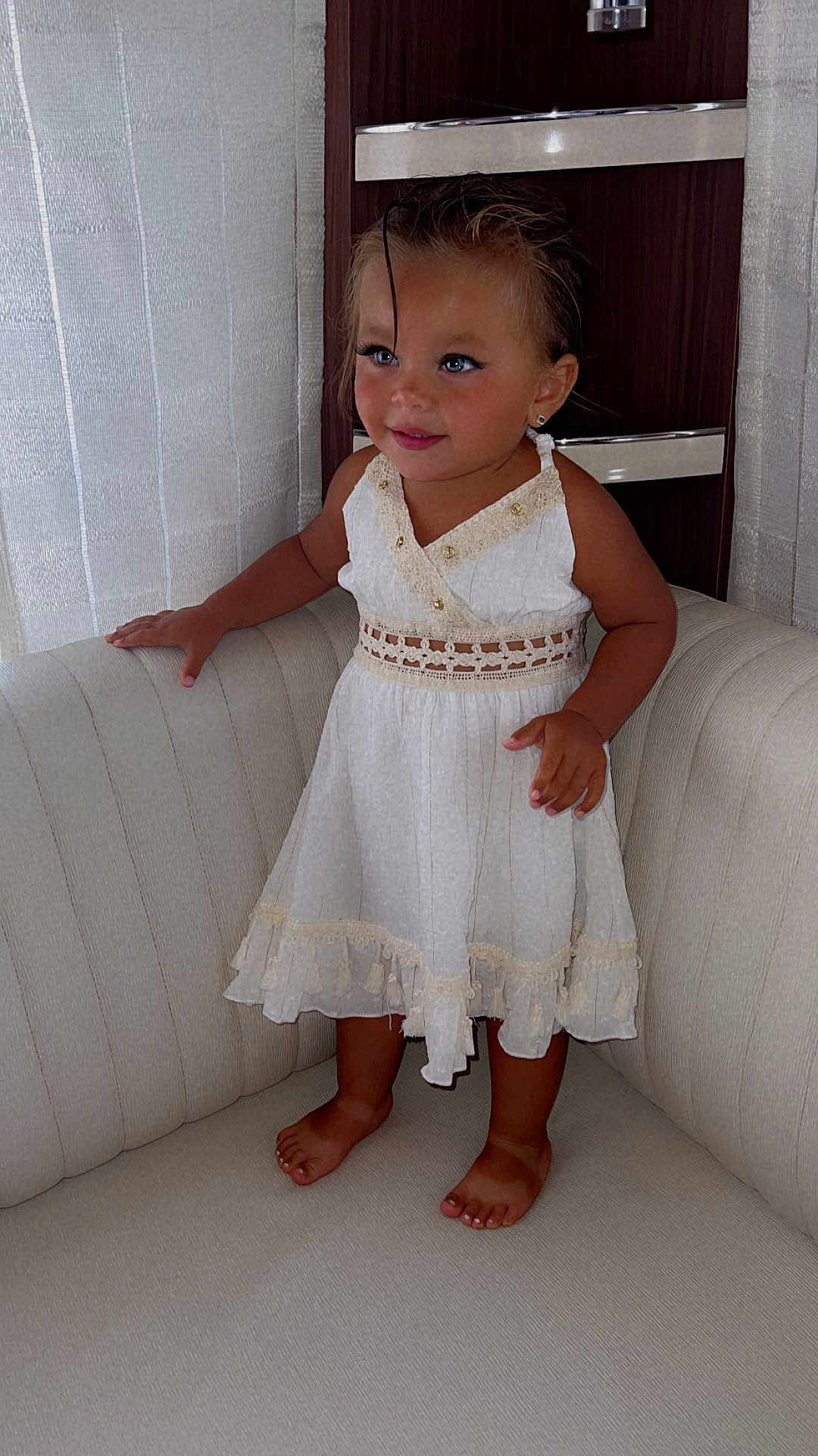 Djaya participe au concours pour gagner de l'argent avec cette photo : toddler, child, barefoot, white_dress, lace, couch, smiling, curly_hair, indoors, furniture, curtains, wooden_background, cute, portrait, standing, happy, small_child, young, person, home