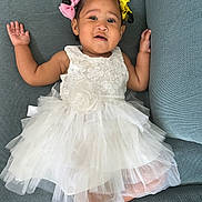 Rayahanna is registered to the contest to win money with this photo: baby, child, dress, white_dress, couch, blue_couch, hair_bows, pink_bow, yellow_bow, smile, face, portrait, cute, infant, sitting, hands_up, tulle, fabric, indoor, person
