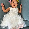 baby, child, dress, white_dress, couch, blue_couch, hair_bows, pink_bow, yellow_bow, smile, face, portrait, cute, infant, sitting, hands_up, tulle, fabric, indoor, person