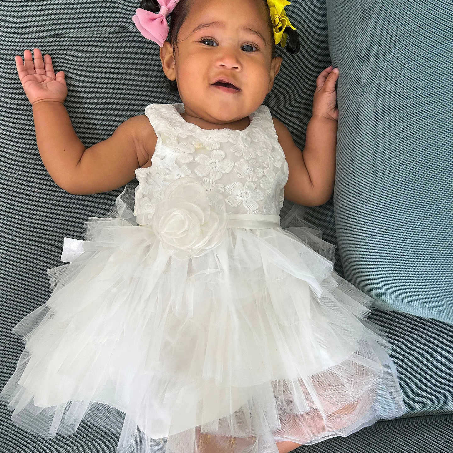 Rayahanna is registered to the contest to win money with this photo: baby, child, dress, white_dress, couch, blue_couch, hair_bows, pink_bow, yellow_bow, smile, face, portrait, cute, infant, sitting, hands_up, tulle, fabric, indoor, person