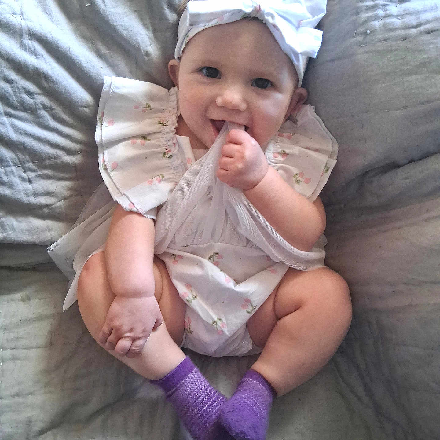 Kinsey is registered to the contest to win money with this photo: baby, child, infant, smiling, happy, sitting, portrait, bow, headband, dress, floral_pattern, purple_socks, finger_in_mouth, bed, bedding, indoors, gray_background, candid, adorable, photography
