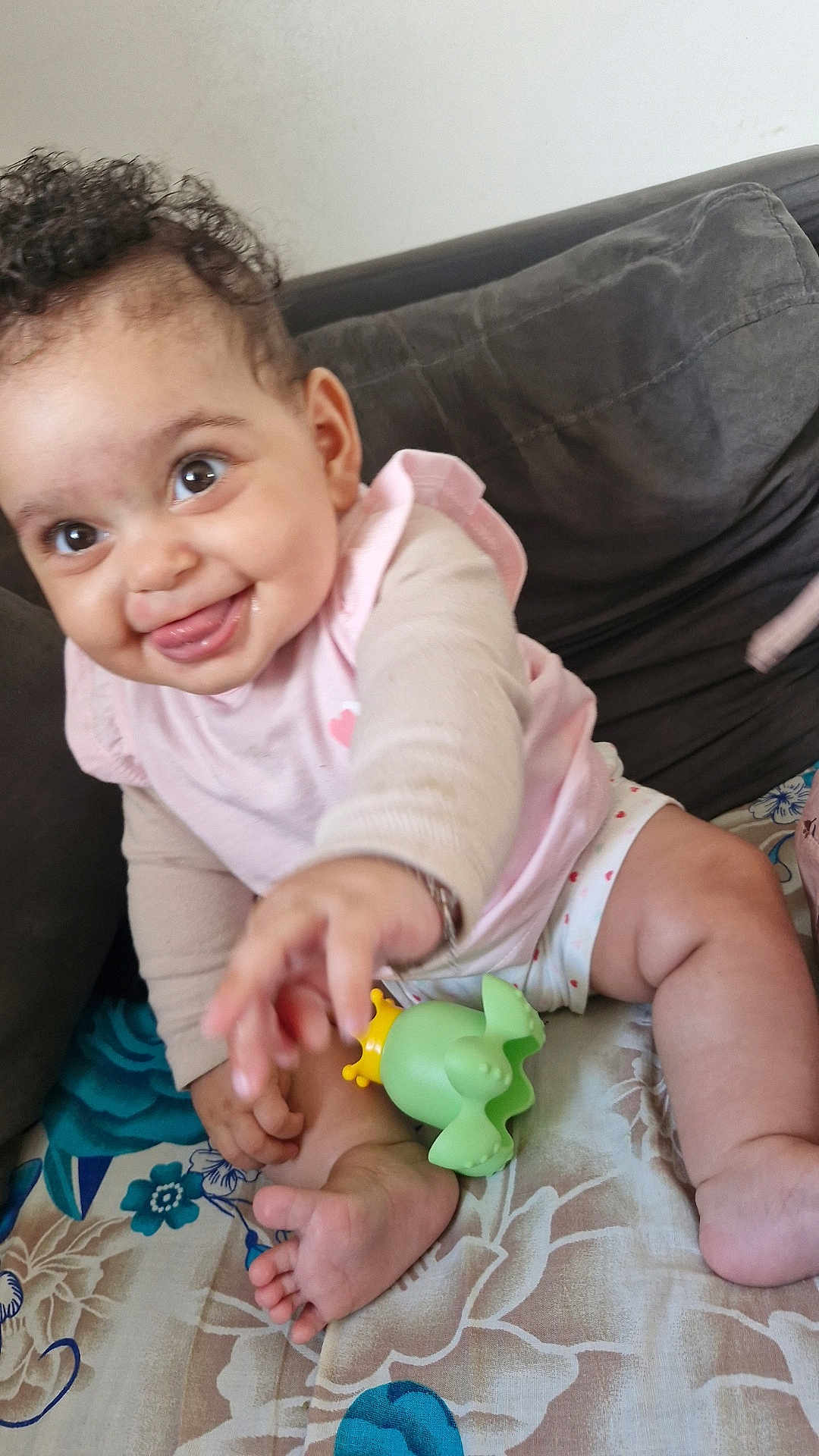 Siyana a rejoint le concours — aidez-le/la à gagner de superbes lots ! baby, child, smiling, tongue_out, curly_hair, toy, couch, floral_pattern, clothing, pink_shirt, barefoot, happy, indoor, person, face, hand, foot, sitting, playful, cute