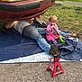 child, toddler, person, adult, car, ford_mustang, vehicle, jack_stand, tool, outdoor, grass, gravel, jeans, pink_clothing, repair, mechanic, play, sitting, working, daylight
