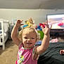 toddler, child, smile, happy, pink_clothing, blonde_hair, indoor, carpet, crib, television, cartoon, toy, person, raising_arms, curly_hair, playful, living_room, cute, expression, home