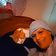 Flannel is registered to the contest to win money with this photo: beanie, bed, blanket, casual_clothing, cat, close_up, cozy, curtain, earrings, facial_expression, indoor, jewelry, orange_cat, person, portrait, relaxed, resting, smile, warm_lighting, white_cat