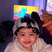 Sajid participe au concours pour gagner de l'argent avec cette photo : cap, cartoon, casual, child, couch, curly_hair, curtain, dinosaur, face, hand, happy, indoor, living_room, person, playful, smiling, sweatshirt, television, toddler, window