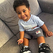 Jayden is registered to the contest to win money with this photo: bright, casual_clothing, child, comfortable, couch, curly_hair, cute, footwear, happy, indoor, living_room, person, portrait, shorts, sitting, smile, sneakers, toddler, tshirt, young_child