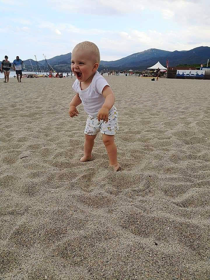 Aloïs a rejoint le concours — aidez-le/la à gagner de superbes lots ! beach, body_of_water, cloud, ecoregion, fun, gesture, grass, happy, horizon, landscape, natural_environment, people_in_nature, people_on_beach, person, sand, shore, shorts, sky, t_shirt, toddler