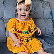 Jimena is registered to the contest to win money with this photo: baby, child, girl, smile, yellow_dress, bow, headband, sitting, couch, indoor, happy, cute, portrait, plush, furniture, embroidery, barefoot, person, young, bright