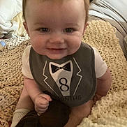 Ethan is registered to the contest to win money with this photo: baby, smiling, bib, tuxedo_design, indoor, blanket, plush_toy, child, cute, sitting, face, head, clothing, soft_lighting, person, infant, cozy, home, young_child, happy
