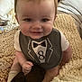 baby, smiling, bib, tuxedo_design, indoor, blanket, plush_toy, child, cute, sitting, face, head, clothing, soft_lighting, person, infant, cozy, home, young_child, happy