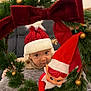 baby, child, red_hat, knit_hat, christmas_wreath, holiday_decor, plush_elf, ornaments, greenery, curious, indoor, festive, cute, face, portrait, seasonal, smiling, soft_lighting, cozy, button_clothing