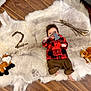 baby, fur_rug, wood_floor, plaid_shirt, brown_pants, toy_axe, plush_fox, plush_squirrel, sticks, number_two, indoor, child, flooring, cute, infant, wood_texture, soft_texture, lying_down, playful, decorative