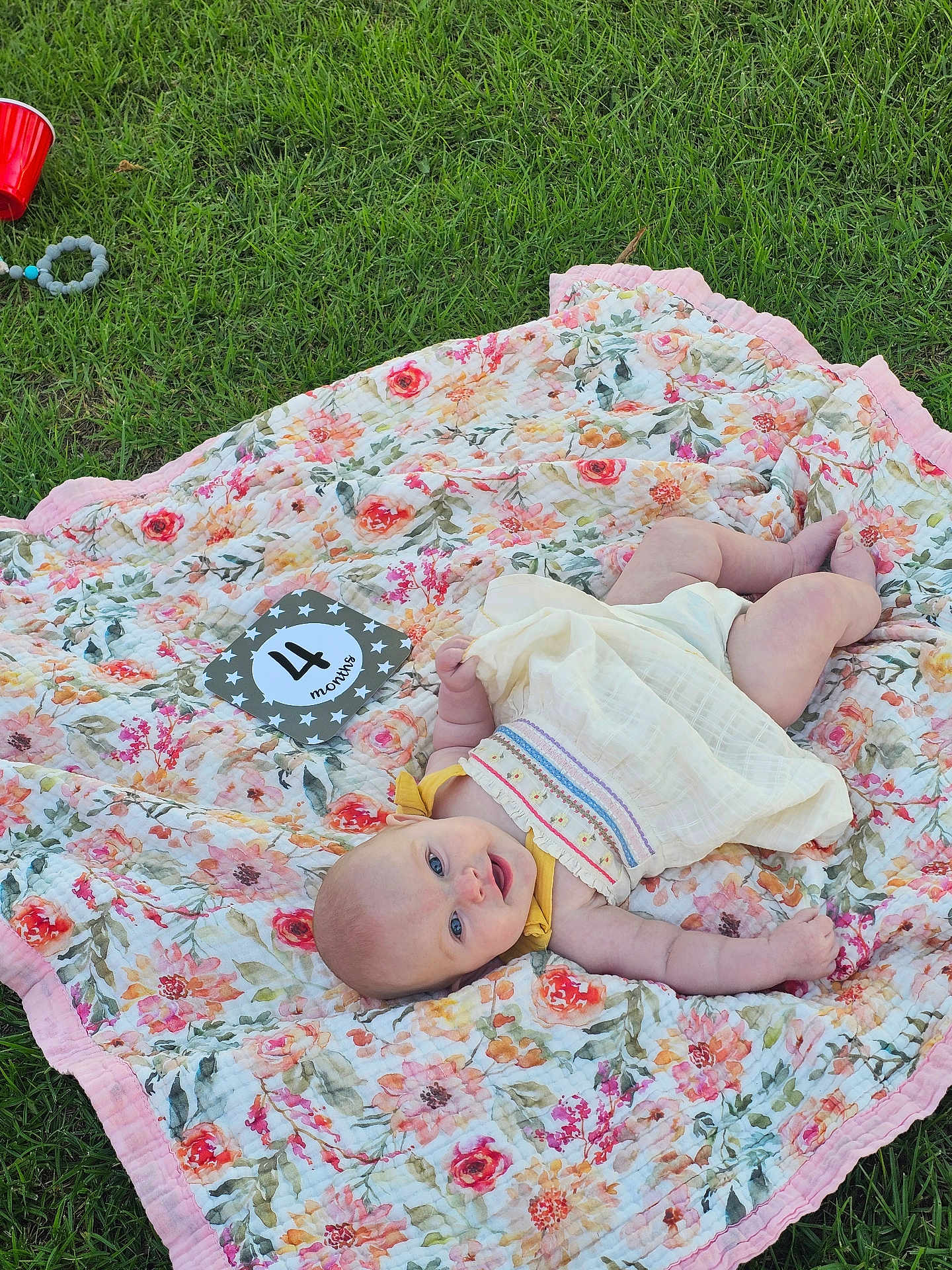 Elodie joined the competition — help win amazing prizes! baby, infant, blanket, floral_pattern, grass, outdoor, smiling, milestone_card, four_months, dress, cute, pacifier, red_cup, summer, relaxing, lying_down, child, nature, happy, portrait