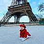 baby, red_coat, red_beret, eiffel_tower, outdoor, sky, cloud, concrete, happy, child, tourist_spot, landmark, architecture, smile, winter_clothing, trees, person, sitting, urban, daytime