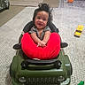 Desi joined the competition — help win amazing prizes! baby, child, smiling, sitting, toy_car, walker, red_pillow, indoor, living_room, rug, carpet, toy_truck, orange_ball, green_toy, gray_shirt, dark_hair, happy, portrait, playtime, household