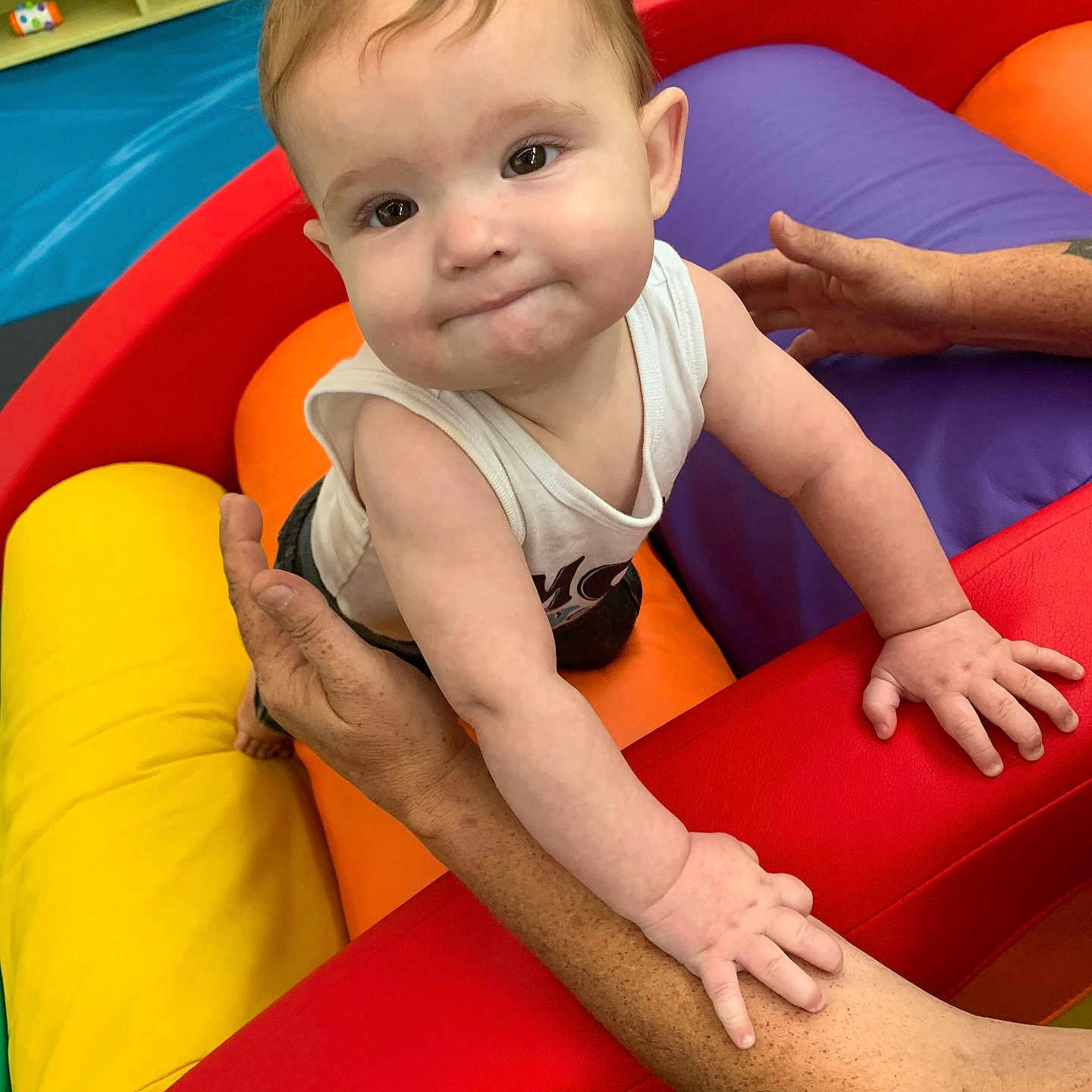 Samuel is registered to the contest to win money with this photo: baby, child, infant, portrait, smiling, determined, face, eyes, hair, white_shirt, ball_pit, colorful, play_area, indoor, toys, foam_balls, red, yellow, purple, adult_hands