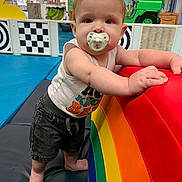 Samuel is registered to the contest to win money with this photo: baby, child, infant, pacifier, standing, indoor, playground, play_area, rainbow, cushion, soft_play, toys, colorful, portrait, face, brown_hair, sleeveless_shirt, shorts, smiling, playroom