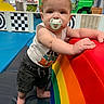 baby, child, infant, pacifier, standing, indoor, playground, play_area, rainbow, cushion, soft_play, toys, colorful, portrait, face, brown_hair, sleeveless_shirt, shorts, smiling, playroom