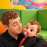 Samuel joined the competition — help win amazing prizes! baby, infant, child, adult, father, parent, cuddle, pacifier, portrait, face, sleeping, wide_eyes, brown_hair, black_clothing, green_couch, colorful, indoor, playground, underwater_mural, cartoon_style