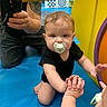 Samuel joined the competition — help win amazing prizes! baby, child, infant, pacifier, crawling, sitting, smiling, face, indoor, play_area, colorful, toy, playground, adult, smartphone, photography, black_clothing, blue_floor, yellow_structure, family