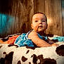 baby, infant, child, blanket, cow_print, blue_clothing, red_shorts, wooden_background, cozy, soft_texture, cute, curious, lying_down, indoors, portrait, warm_lighting, skin, face, arms, expression