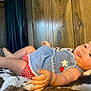baby, infant, tongue_out, blue_top, red_checkered_shorts, blanket, wooden_wall, curtain, painting, indoor, child, cute, playful, skin, legs, hand, face, expression, cozy, relaxed