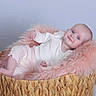 Lila is registered to the contest to win money with this photo: baby, infant, basket, pink_fur, white_clothing, smiling, face, feet, hand, floor, wooden_floor, woven_basket, portrait, indoor, cozy, cute, resting, child, soft_texture, relaxed