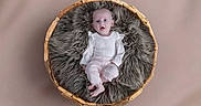 Lila is registered to the contest to win money with this photo: baby, infant, basket, fur, soft, cozy, white_clothing, pink_pants, floor, neutral_background, portrait, indoor, cute, child, small, sitting, looking_up, expression, comfortable, furniture
