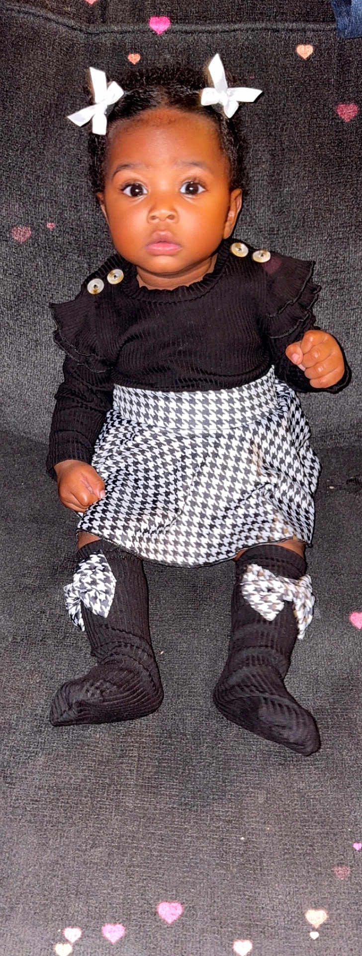 Audree joined the competition — help win amazing prizes! baby, child, toddler, black_clothing, houndstooth_pattern, bows, socks, sitting, indoor, curious_expression, button_details, face, person, cute, portrait, patterned_skirt, dark_background, heart_shapes, fashion, young_child