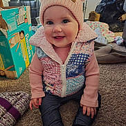Elizabeth is registered to the contest to win money with this photo: baby, child, pink_hat, pom_poms, patchwork_vest, carpet, indoor, smiling, sitting, pants, long_sleeve, toy_box, pampers_box, household_items, footwear, cute, portrait, face, person, cozy