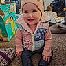 baby, child, pink_hat, pom_poms, patchwork_vest, carpet, indoor, smiling, sitting, pants, long_sleeve, toy_box, pampers_box, household_items, footwear, cute, portrait, face, person, cozy