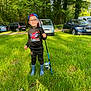 child, kid, person, glasses, cap, backwards_cap, fishing_rod, fish, rain_boots, spiderman_sweatshirt, grass, park, trees, cars, parking, outdoors, smile, sunlight, nature, portrait