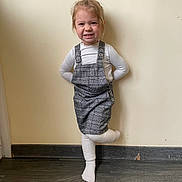 Lyna participe au concours pour gagner de l'argent avec cette photo : child, toddler, girl, smile, plaid_pinafore, white_top, tights, socks, standing, one_leg, indoor, plain_wall, hardwood_floor, portrait, hands_behind_back, messy_hair, playful, cute, full_body, young